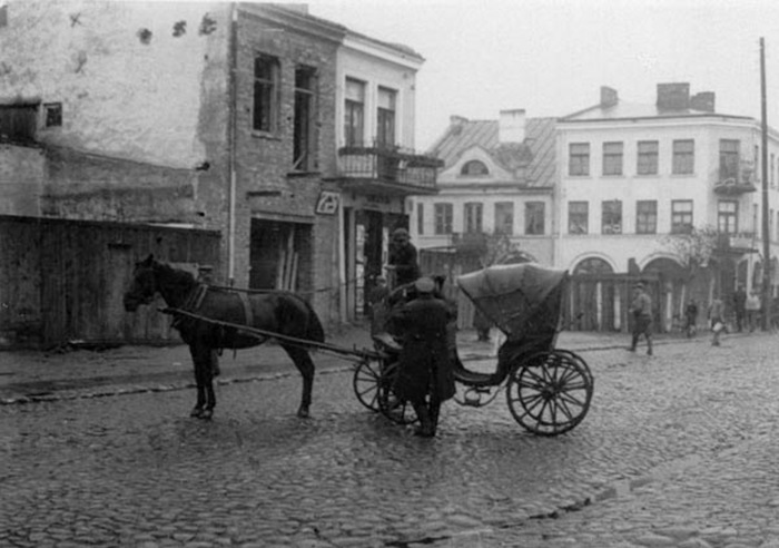 Zdjęcie przedstawia dorożkę zaprzężoną w konie na ulicy. Przed dorożką stiu mężczyzna