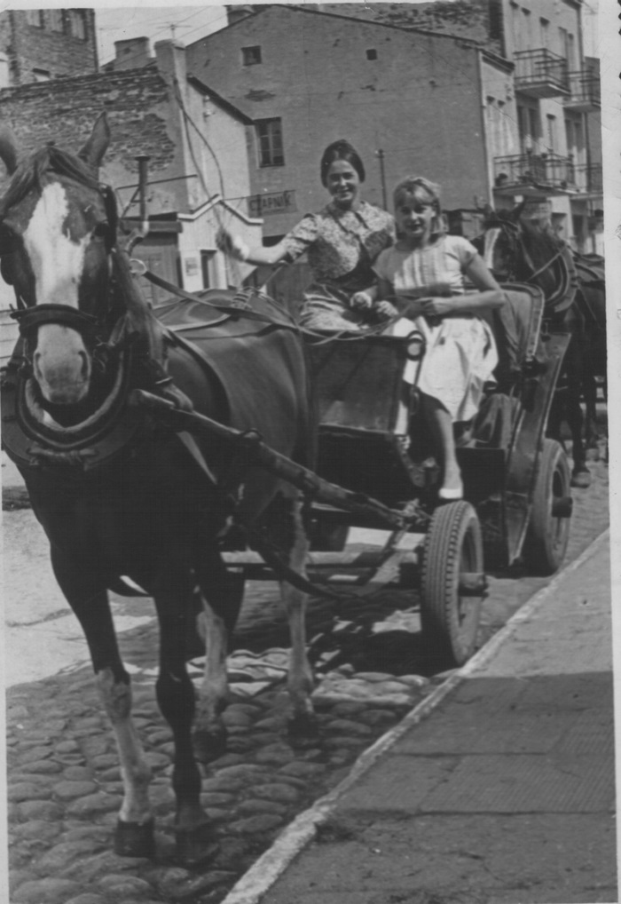 Zdjęcie przedstawia dwie kobiety jadące dorożką, na pierwszym planie głowa konia