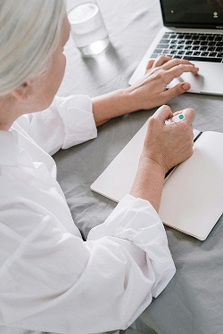 zdjęcie przedstawia starszą kobietę pracującą przy laptopie i robiocą notatki