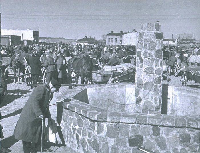 Zdjęcie przedstawia starszego mężczyznę przy kamiennej fontannie. W tle odbywa się targ - widać konie, wozy i&nbsp;wielu ludzi