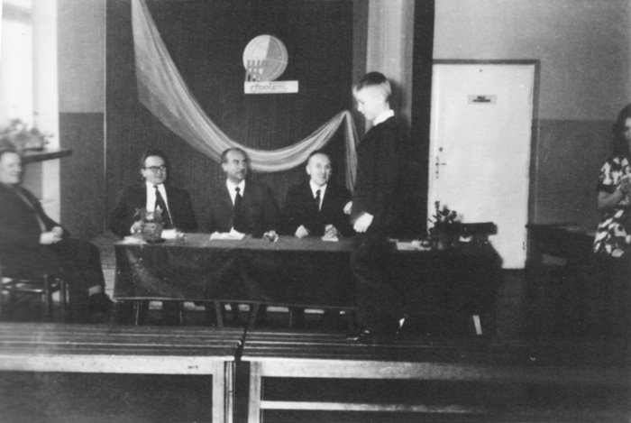 zdjęcie przedstawia chłopca idącego po wybiegu, za nim przy stole siedzi czterech mężczyzn w&nbsp;garniturach