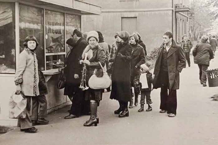 Czarno-białe zdjęcie przedstawia grupę ludzi stojących przy kiosku z&nbsp;gazetami