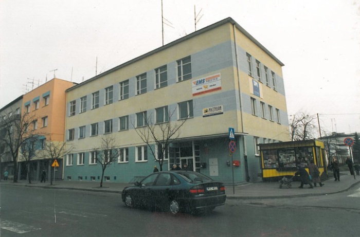 zdjęcie przedstawia widok na&nbsp;ulicę i&nbsp;stojące przy niej budynki, w&nbsp;tym mały ż&oacute;łty kiosk. Na pierwszym planie ciemny samoch&oacute;d