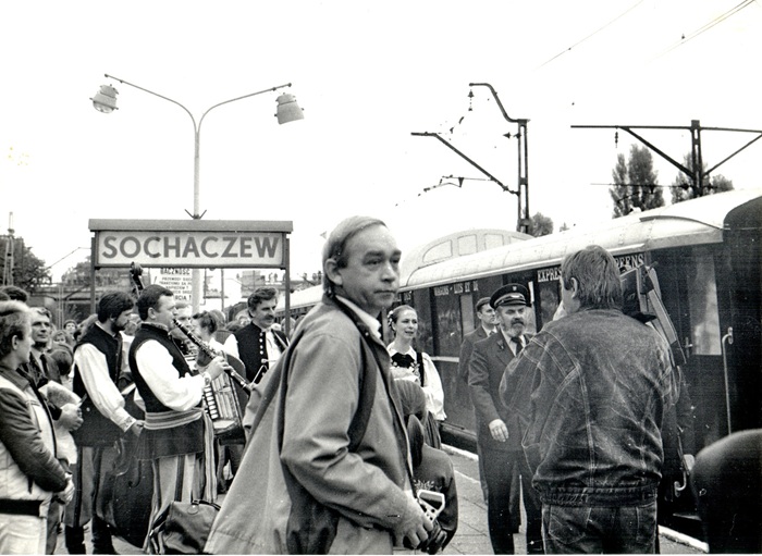 czarno-białe zdjęcie przedstawia pociąg na&nbsp;peronie, dużo os&oacute;b, na&nbsp;pierwszym planie mężczyzna a&nbsp;aparatem fotograficznym